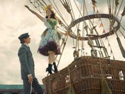 A young white man in a suit stands at the foot of a hot air balloon as a young white woman in a colourful dress stands on the basket with her arm in the air