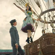 A young white man in a suit stands at the foot of a hot air balloon as a young white woman in a colourful dress stands on the basket with her arm in the air
