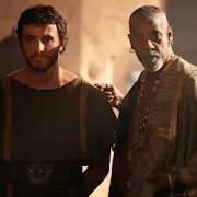 A young white man in plain robes with his hands behind his back stands besides a middle aged Black man in elegant robes