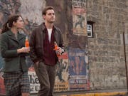 A man and a woman walking away from a brick wall full of wartime posters, holding fizzy drink bottles. I