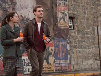 A man and a woman walking away from a brick wall full of wartime posters, holding fizzy drink bottles. I