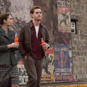 A man and a woman walking away from a brick wall full of wartime posters, holding fizzy drink bottles. I