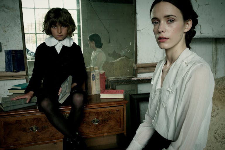 A woman in a white shirt and creepy child in a black and white 1800 school uniform are looking intensely at the camera