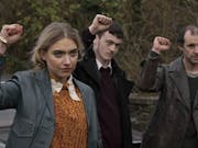 A woman and two men raising their fists are looking determinedly at something or someone on the right of the camera