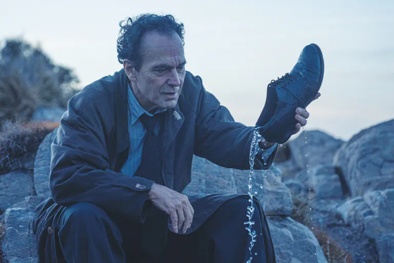 A white man sitting on some rocks is pouring water out of his shoe