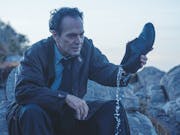A white man sitting on some rocks is pouring water out of his shoe