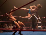 In a boxing ring, a man is in the air having just landed a kick in the head of his opponent who is leaning backwards