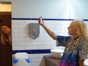A blonde woman in public toilets is videoing herself with her phone while another blonde woman peeks from the entrance and looks at her.