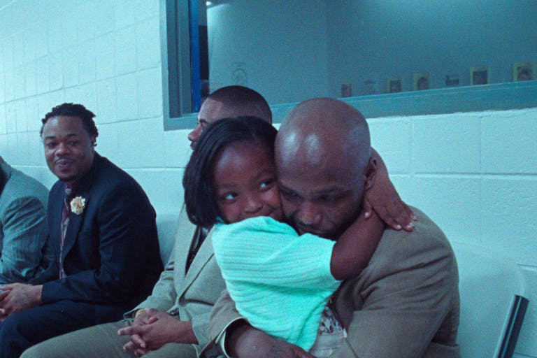 A little Black girl hugs a Black man in a white room with a mirror behind them, while other people look on and smile.