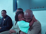 A little Black girl hugs a Black man in a white room with a mirror behind them, while other people look on and smile.