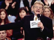White man holding cue cards that read Jerry Springer' is speaking to the camera while a large crow
