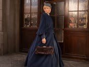 A white woman in a large Victorian blue dress and a hat walks in front of a building with glass doors
