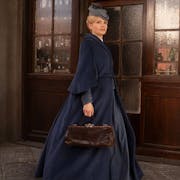 A white woman in a large Victorian blue dress and a hat walks in front of a building with glass doors