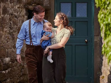 A white man and a woman in front of a house are smiling at each other. The woman is holding a baby