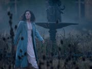 A woman in a dress and long coat is walking in front of a dry fountain. Around her dead flowers.