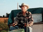 An elderly man with a large moustache wearing jeans a check shirt and a cowboy hat is driving a tractor with