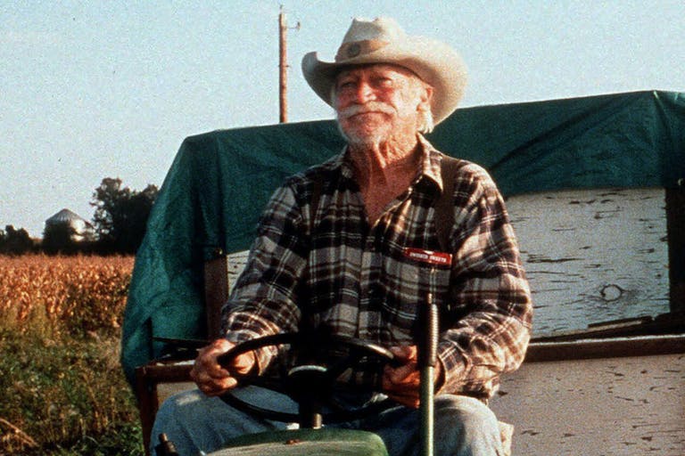 An elderly man with a large moustache wearing jeans a check shirt and a cowboy hat is driving a tractor with