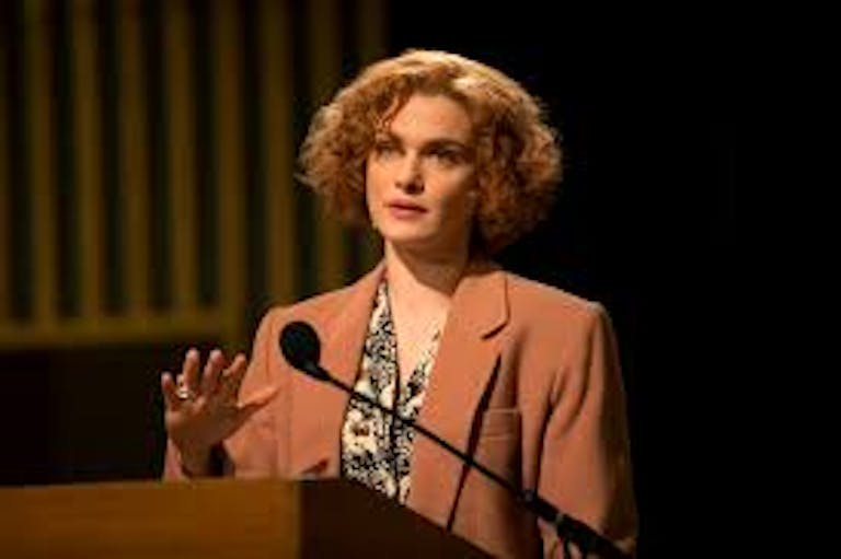 White woman with short curly hair is standing on a podium giving a speech