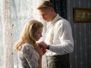 A blond man and blonde woman in 1940s clothes are standing by a window facing each other looking concerned and holding hands.