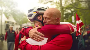 A man with shaved head and one wearing cycling gear and helmet are embracing in a leafy street full of people and flags