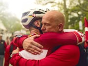 A man with shaved head and one wearing cycling gear and helmet are embracing in a leafy street full of people and flags