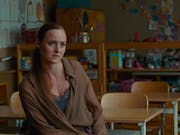 A young white woman with long tied up hair and loose brown shirt sits in a classroom facing the teacher's desk looking serious