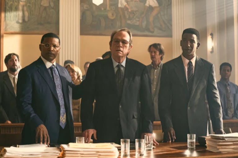 Two Black men and a middle aged white man stand behind a desk with papers piled in a courtroom