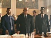 Two Black men and a middle aged white man stand behind a desk with papers piled in a courtroom