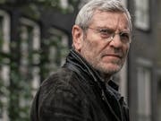 An older white man with grey hair, glasses and a dark jacket looking serious