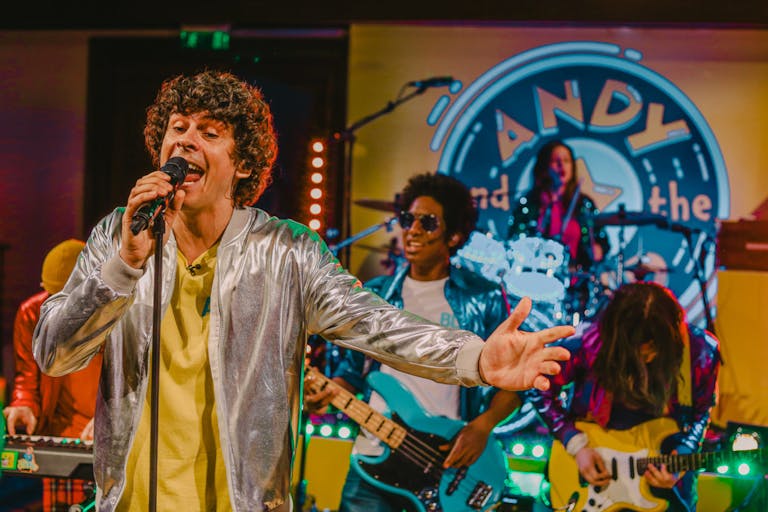 Five men and white in shinny costumes playing various instruments and singing, performing as a band