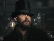 A white man with a rugged beard wearing a tall hat, gas lamps in the background