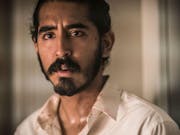A young British-Indian man with dark facial hair wearing a loose white shirt looking dishevelled and concerned