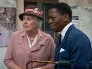 A young Black man in a blue suit stands talking to an old white woman in a pink coat and hat looking thoughtful