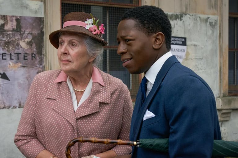A young Black man in a blue suit stands talking to an old white woman in a pink coat and hat looking thoughtful