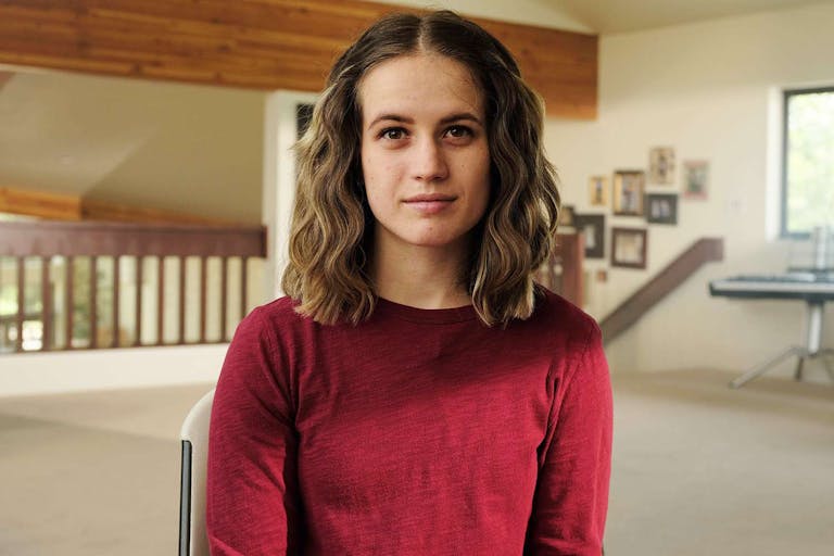 A young white woman with short brown hair wearing a red top sitting giving a talking head