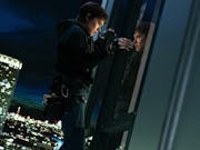 At night, a white woman with short brown hair climbs a tall glass fronted building