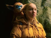 A young blonde white girl in yellow puffer jacket looking amazed with a small blue faced, brown furred monkey-type creature with large ears sits on her shoulder