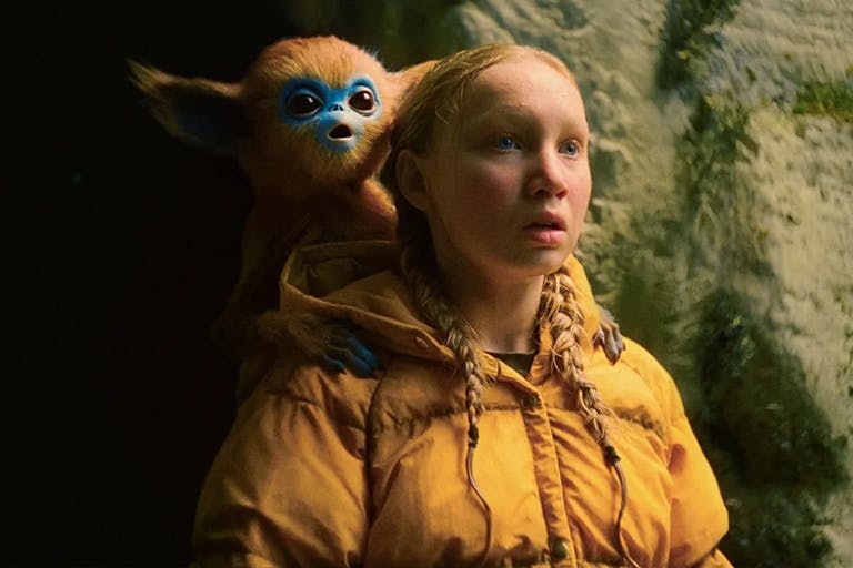 A young blonde white girl in yellow puffer jacket looking amazed with a small blue faced, brown furred monkey-type creature with large ears sits on her shoulder