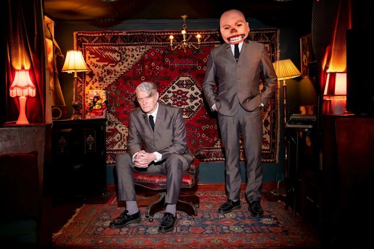 Two white men in dark grey suits in a lamp-lit room with red furnishings, one of the men's faces is photoshopped, enlarged and distorted