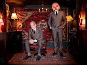 Two white men in dark grey suits in a lamp-lit room with red furnishings, one of the men's faces is photoshopped, enlarged and distorted