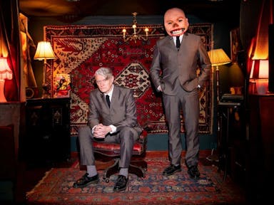Two white men in dark grey suits in a lamp-lit room with red furnishings, one of the men's faces is photoshopped, enlarged and distorted