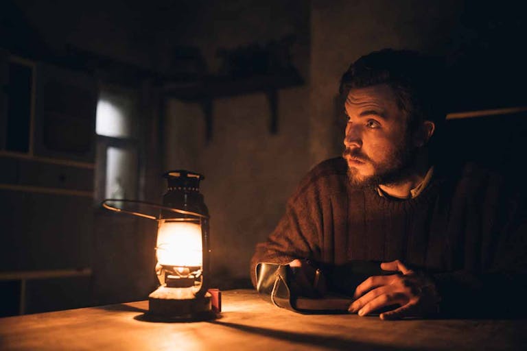 A young white man in a thick jumper sit in a dark room lit up by the orange glow of a lantern