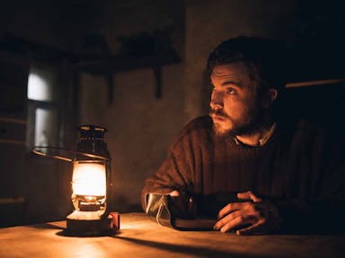 A young white man in a thick jumper sit in a dark room lit up by the orange glow of a lantern