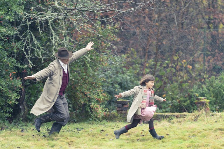 A white man in trench coat and hat, arms outstretched, playing airplanes in the garden with his daughter, arms also outstretched