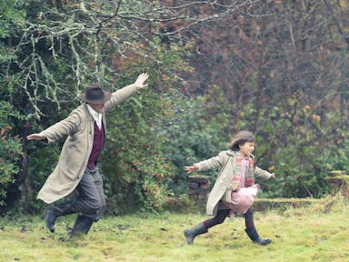 A white man in trench coat and hat, arms outstretched, playing airplanes in the garden with his daughter, arms also outstretched