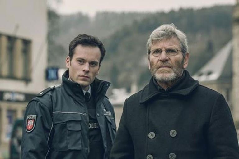 A young white man in foreign police uniform stands next to an older white man with grey hair, glasses and a dark jacket