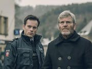 A young white man in foreign police uniform stands next to an older white man with grey hair, glasses and a dark jacket