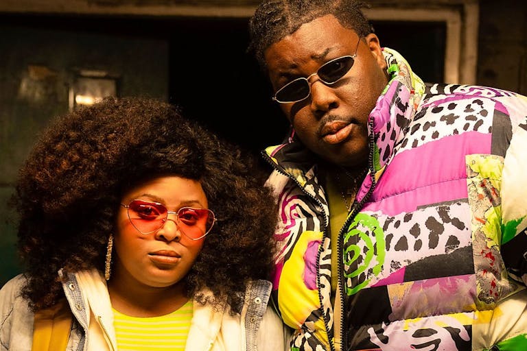 A young Black woman and man in colourful clothing and sunglasses looking seriously at the camera