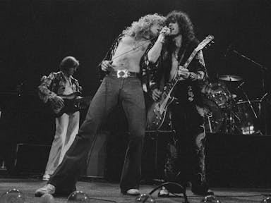 Black and white still of three white men with big hair and 80s clothing on stage playing instruments