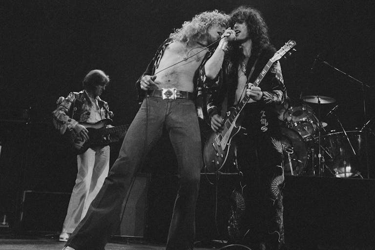 Black and white still of three white men with big hair and 80s clothing on stage playing instruments